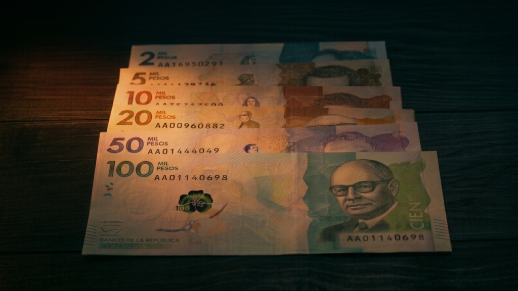 Billetes de pesos colombianos de varias denominaciones sobre superficie oscura, iluminados por luz naranja-ámbar dramática.