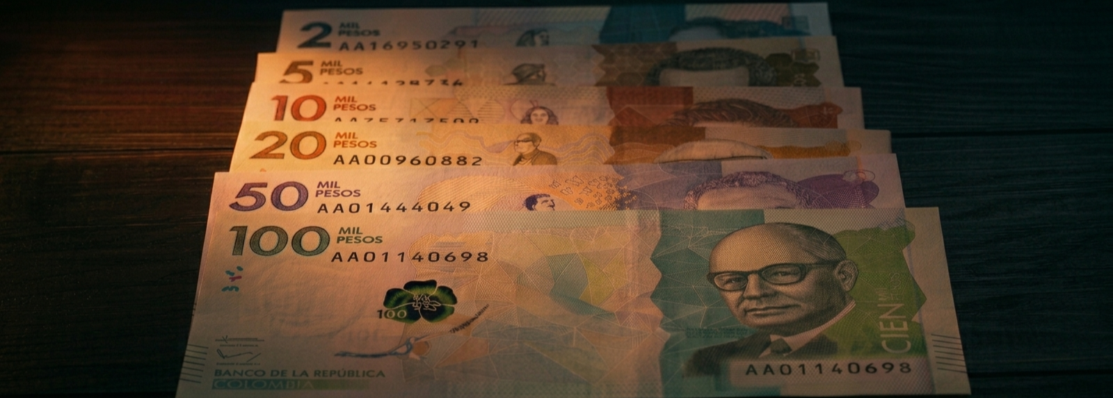 Billetes de pesos colombianos de varias denominaciones sobre superficie oscura, iluminados por luz naranja-ámbar dramática.