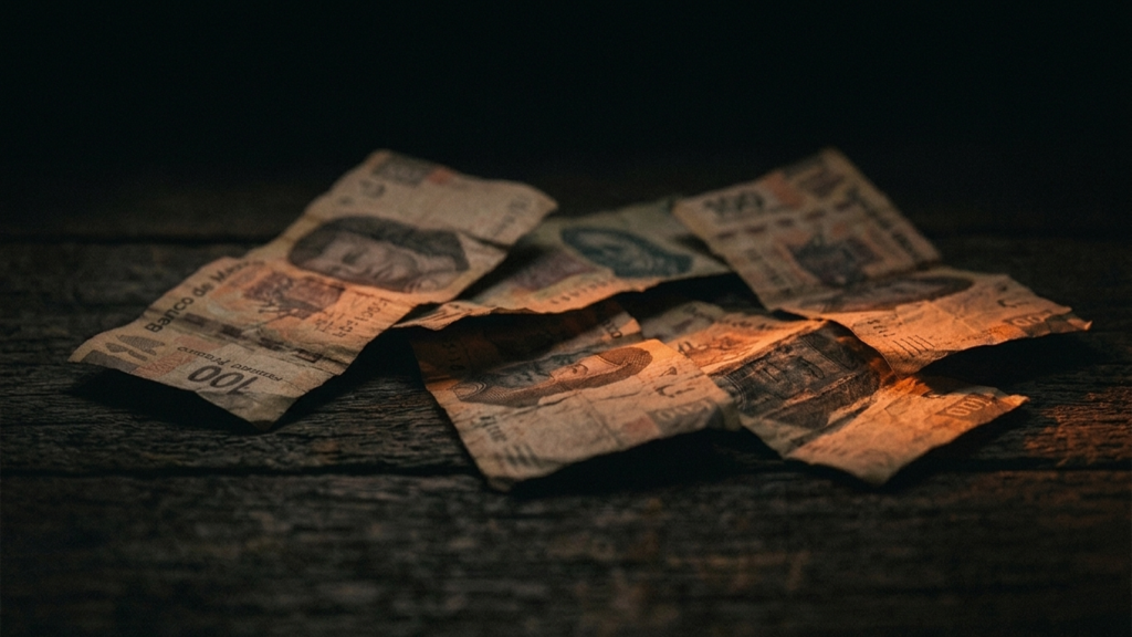 Billetes de peso mexicano arrugados y desgastados sobre una superficie de madera oscura, iluminados por luz naranja-ámbar dramática.
