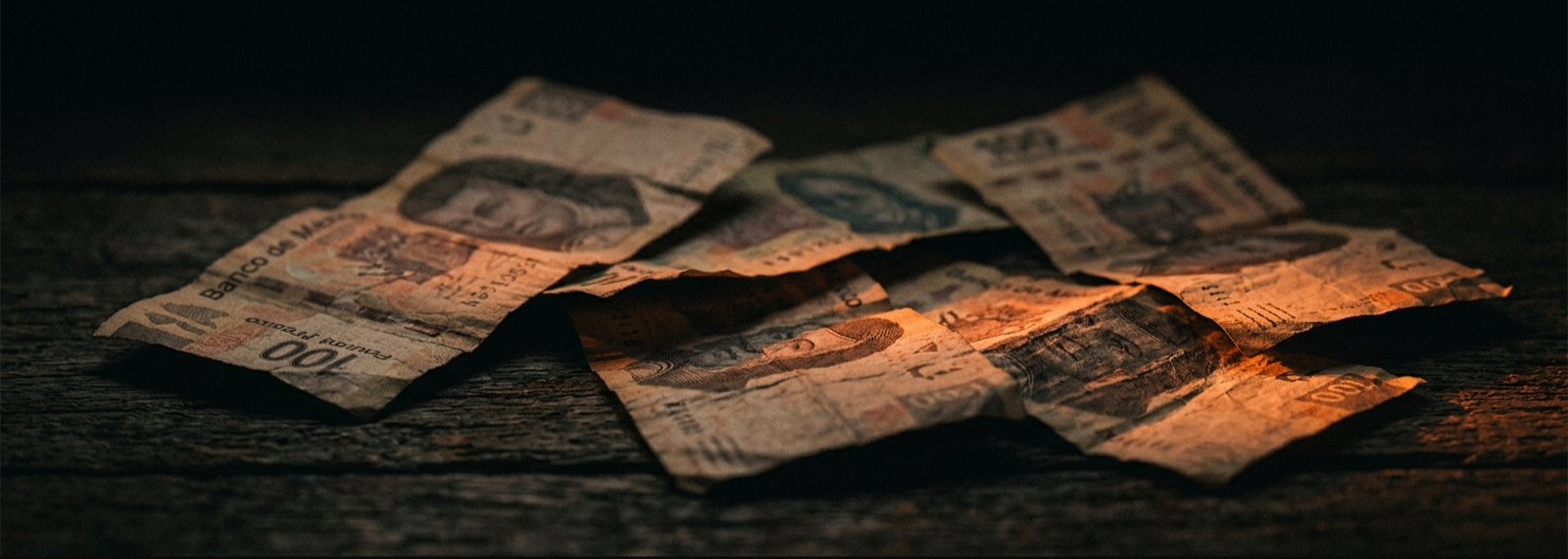 Billetes de peso mexicano arrugados y desgastados sobre una superficie de madera oscura, iluminados por luz naranja-ámbar dramática.