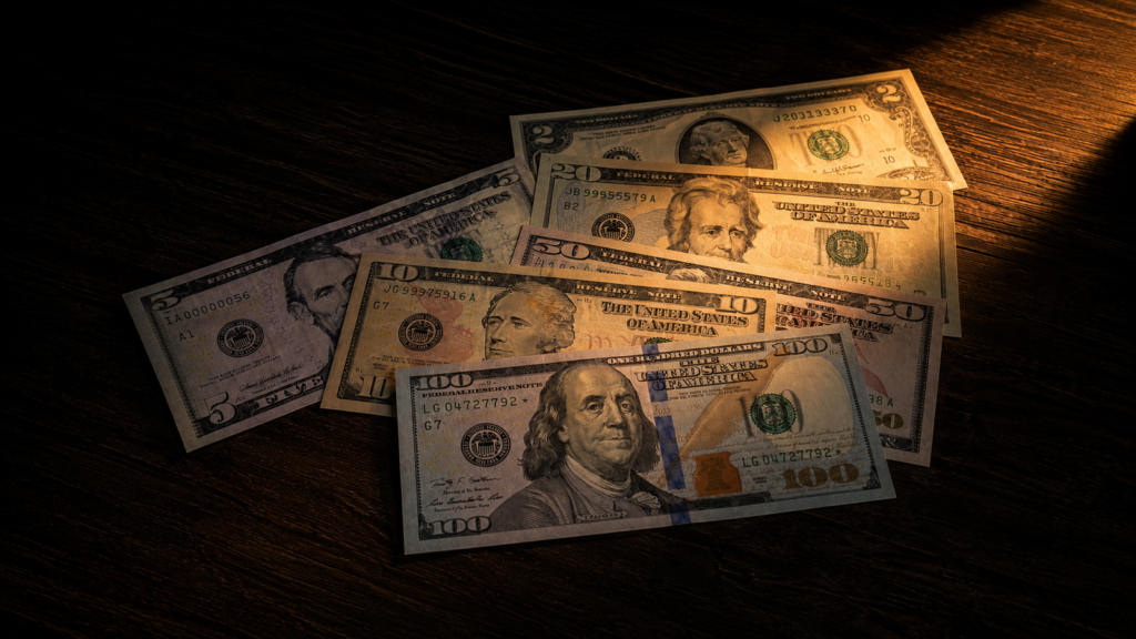 Alt text: Billetes de dólares estadounidenses de varias denominaciones ($5, $10, $20, $50, $100) fanned sobre una mesa de madera oscura, iluminados por una luz naranja-ámbar dramática que recorre el encuadre.